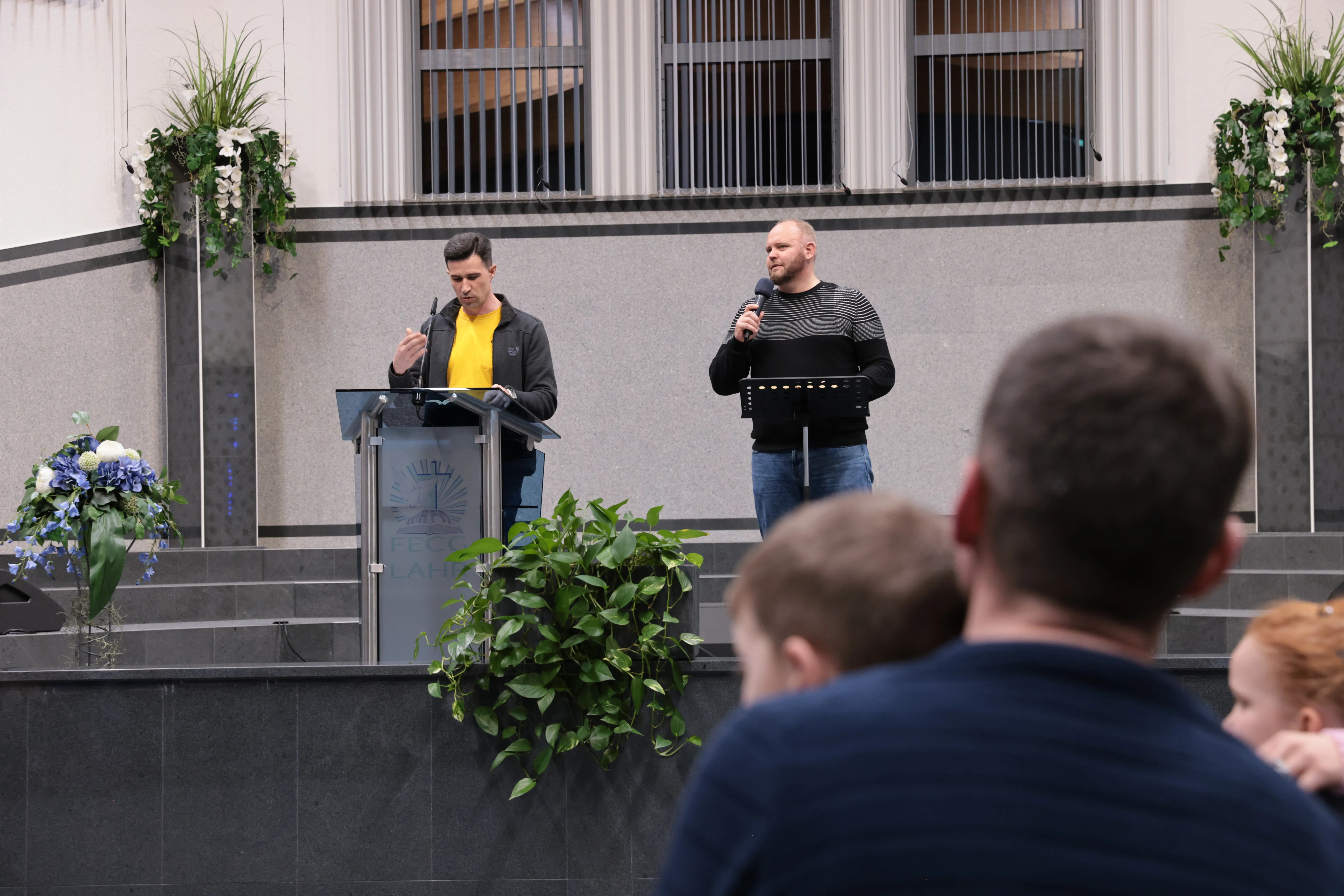 Zwei junge Männer leiten einen Dienst auf der Bühne des Gemeindesaals